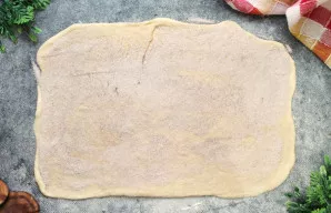 Sumaišykite cukrų ir maltą cinamoną. Darbo paviršių pabarstykite miltais ir iškočiokite tešlą į 42 x 45 centimetrų dydžio stačiakampį. Ap...