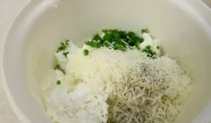 Į atskirą indą sudėkite varškę, tarkuotą parmezaną ir smulkiai supjaustytus svogūnų laiškus. Įpilkite grietinėlės, druskos ir pipirų.