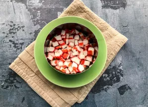 Paimkite serviravimo žiedą ir padėkite jį ant lėkštės. Krabų lazdelės smulkiai supjaustykite ir sudėkite kaip apatinį sluoksnį.