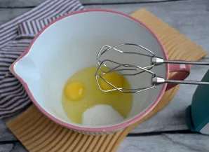 Į dubenį įmuškite 2 kiaušinius, suberkite 60 g cukraus ir išplakite iki putų.